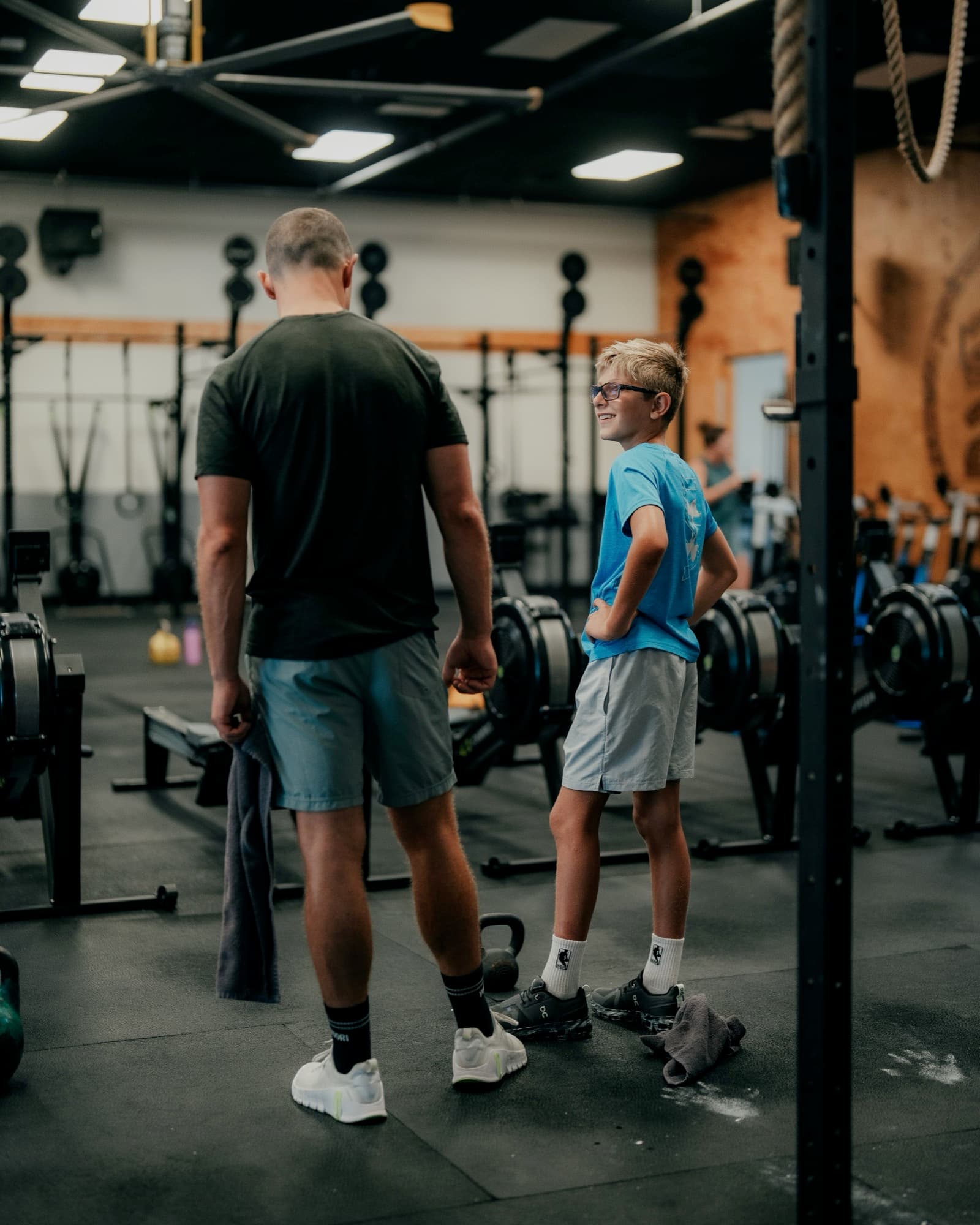 A dad and his son, about eleven, standing together on the gym floor at 2020FIT — the boy, hands on his hips, glancing back up at his dad with a grin, rowing machines and weight racks behind them